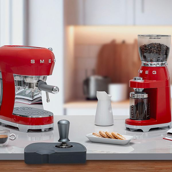 A Smeg retro coffee set including an Espresso Coffee Machine and a Coffee Grinder, surrounded by other coffee accesories including mugs, a carafe, tamping set and decorative cook book and biscuits.