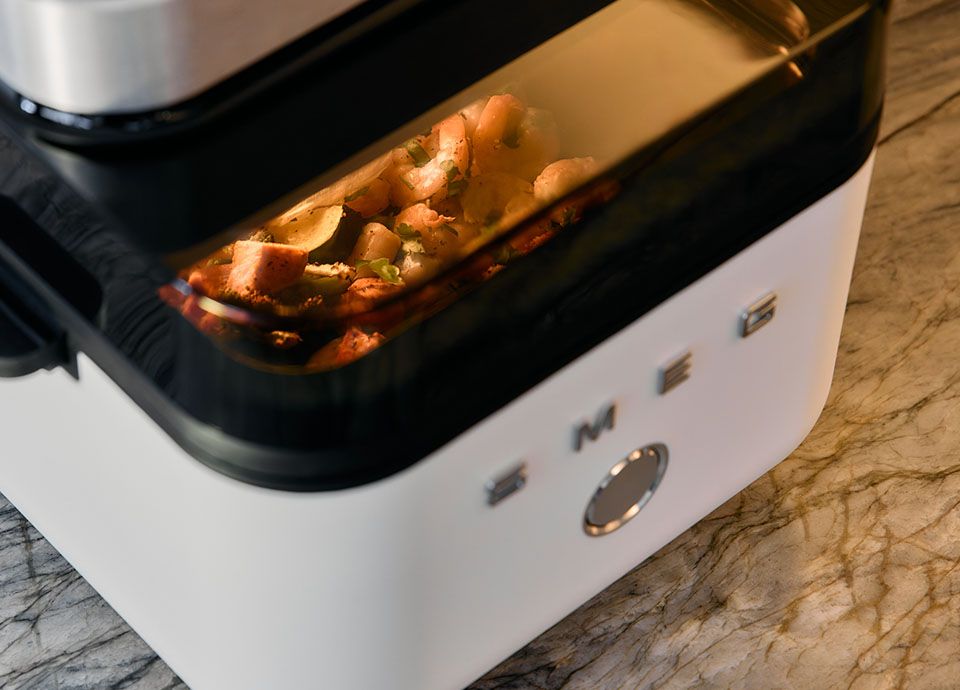 Smeg air fryer in blue finish with transparent window showing muffins, surrounded by eggs, berries and ingredients