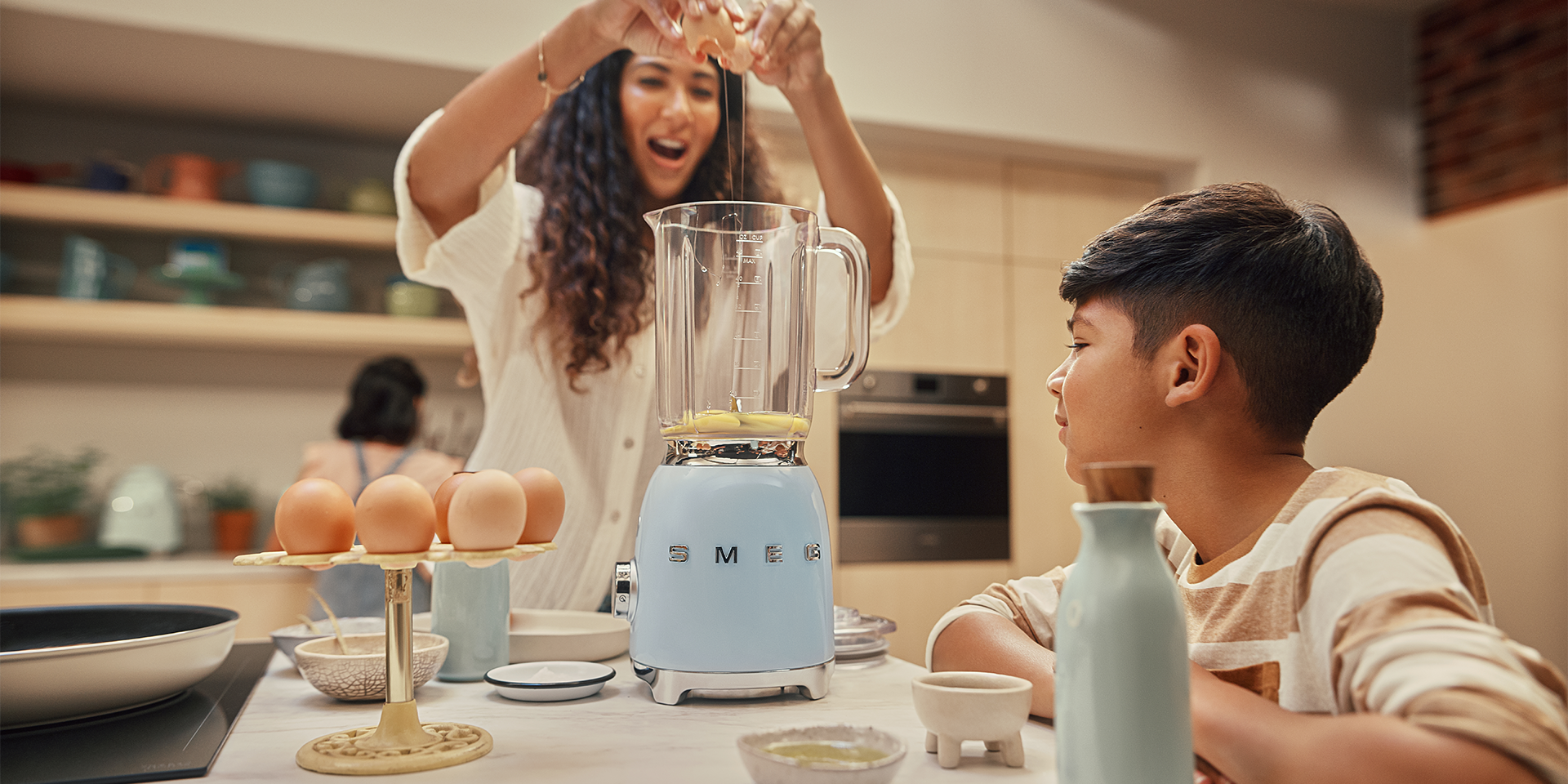 Mother cracking an egg into a Smeg Pastel Blue Blender with a boy watching on