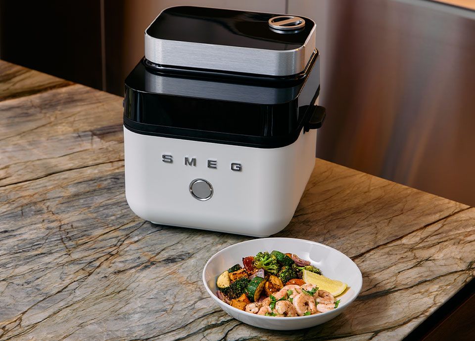 Smeg white air fryer on a wooden countertop surrounded by a variety of dishes including vegetables, chicken and cookies