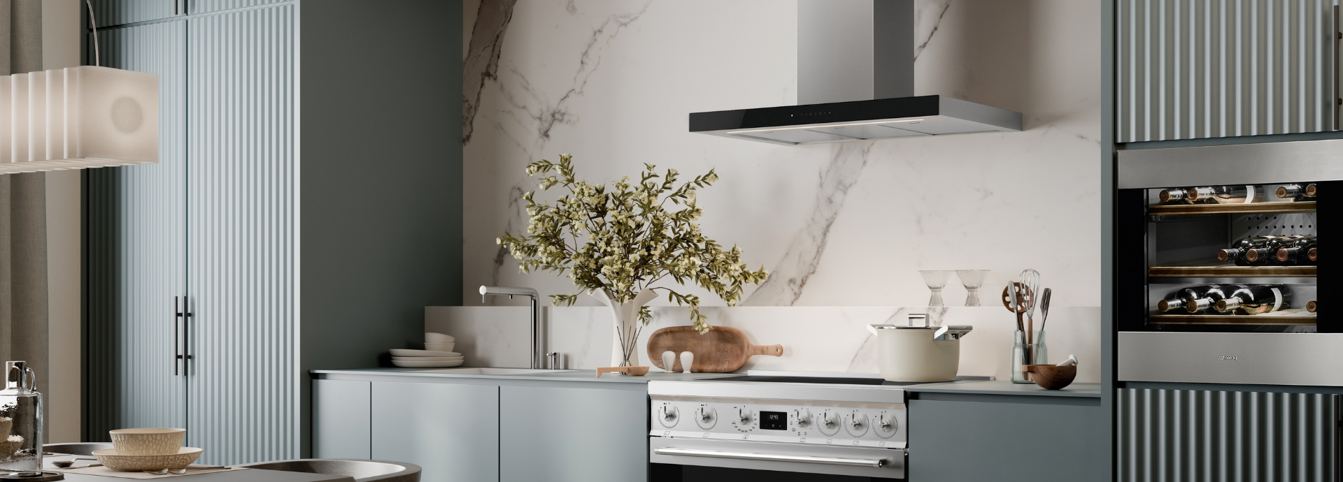 Cuisine moderne et élégante avec électroménagers Smeg intégrés, composée d’un plan de cuisson avec hotte inox suspendue, d’un évier avec robinet chromé et d’un four encastrable  inox avec commandes frontales. Le plan de travail et la crédence en marbre blanc contrastent avec les façades vert d'eau . Des éléments décoratifs en bois, de la vaisselle et un vase de feuillage.