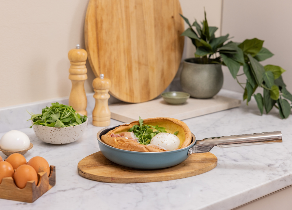Poêle Smeg vert émeraude avec poignée en acier inoxydable. Présentée sur un plan de travail en marbre blanc avec un plateau en bois placé en dessous. La poêle est entourée d’ingrédients et d’ustensiles de cuisine. Une plante verte et des accessoires de cuisine apparaissent en arrière-plan. Mise en scène dans une cuisine moderne.
