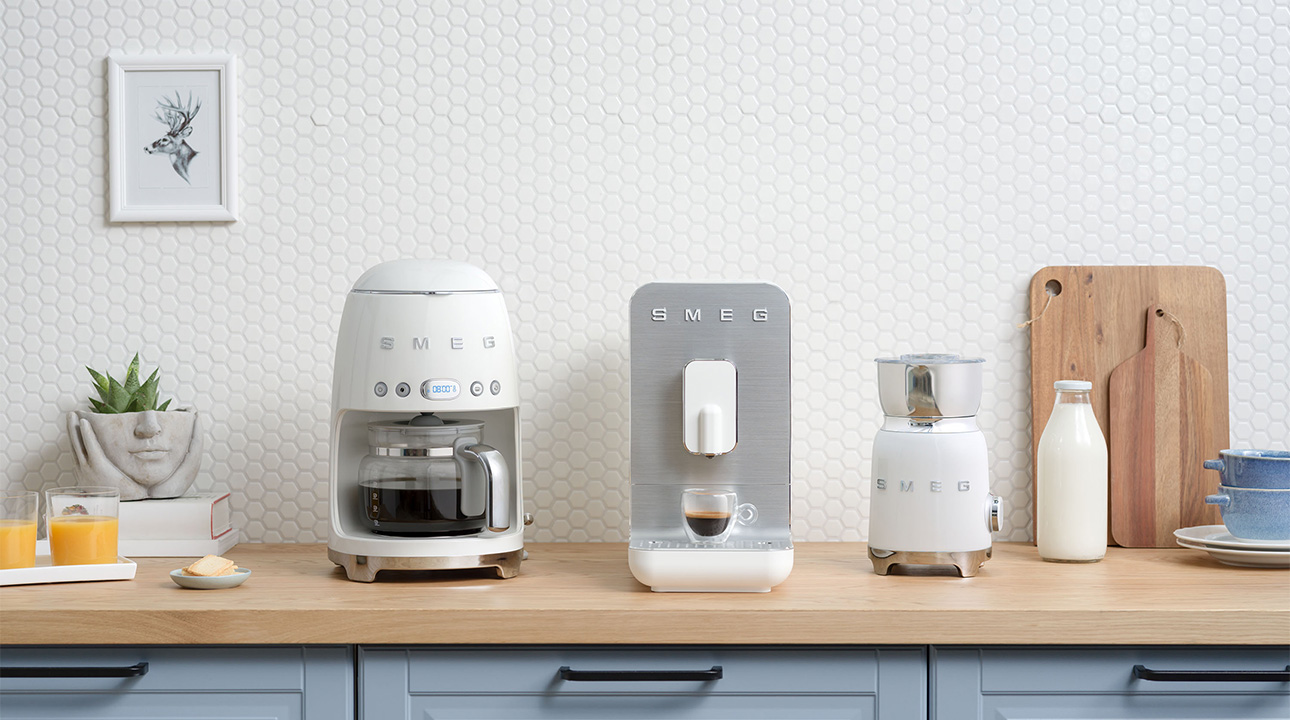 Ensemble café SMEG machine à café automatique blanche, machine à café filtre blanche et mousseur à lait blanc sur un plan de travail.