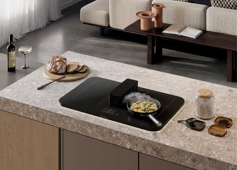 Poêle noire avec poignée en acier inoxydable posée sur une plaque de cuisson à induction. De la fumée s’échappe de la poêle. L’ensemble est installé sur un plan de travail de cuisine où se trouvent du pain, des épices et une bouteille de vin. En arrière-plan, on aperçoit une table basse en bois ainsi que le dossier d’un canapé gris clair.