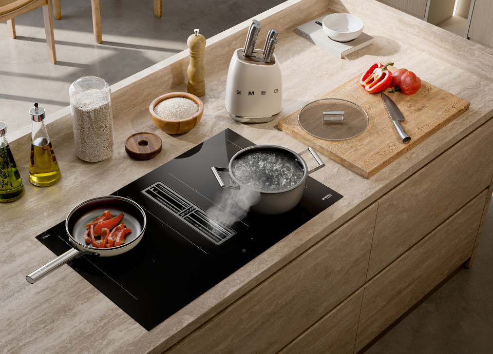 Ensemble de batterie de cuisine Smeg composé d’une casserole et d’une poêle. De l’eau est en train de bouillir dans la casserole, tandis que des poivrons cuisent dans la poêle. Les deux ustensiles sont posés sur une plaque de cuisson à induction avec hotte intégrée. L’ensemble est installé sur un plan de travail en bois avec des tiroirs. Autour, on trouve divers ustensiles de cuisine, un bloc de couteaux blanc Smeg et plusieurs épices.