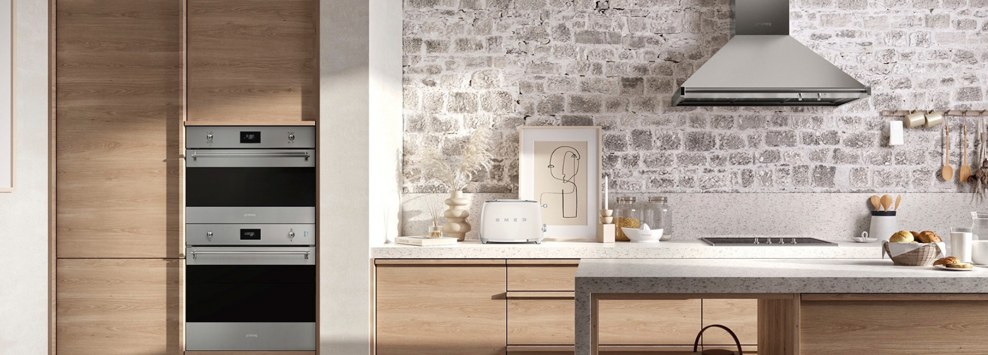 Fours encastrables Smeg esthétique Classica avec finition inox et verre Noir Eclipse, intégrés dans une colonne de meubles en bois naturel au sein d’une cuisine lumineuse. Dotés de poignées tubulaires métalliques, de manettes de commande chromées et d’afficheurs numériques discrets, les fours constituent l’élément principal de la composition. À proximité, une hotte décorative murale en inox de la même collection est installée au-dessus d’une table de cuisson intégrée dans un plan de travail clair. L’espace est complété par un grille-pain Smeg assorti, des accessoires décoratifs, une crédence en briques apparentes et un îlot central aux lignes épurées.