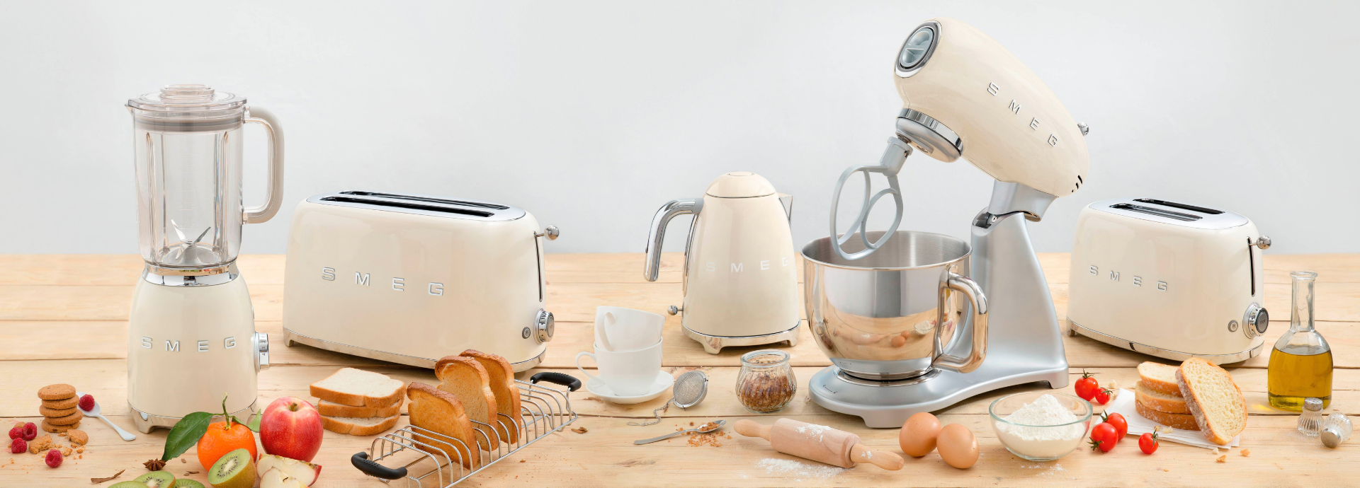 Collection de petit électroménager Smeg composée d’un blender, d'un grille-pain avec ses pinces, d’une bouilloire et d’un robot sur socle avec bol en inox, présentés dans une finition crème au style années 50. Les appareils sont disposés sur une table en bois entourée d’ingrédients et d’ustensiles de cuisine, tels que des tranches de pain, de la farine, des œufs, des fruits frais, de l’huile d’olive, une tasse à café et un rouleau à pâtisserie. L’ensemble est mis en scène sur un fond épuré clair mettant en valeur le design rétro et les finitions chromées des appareils.