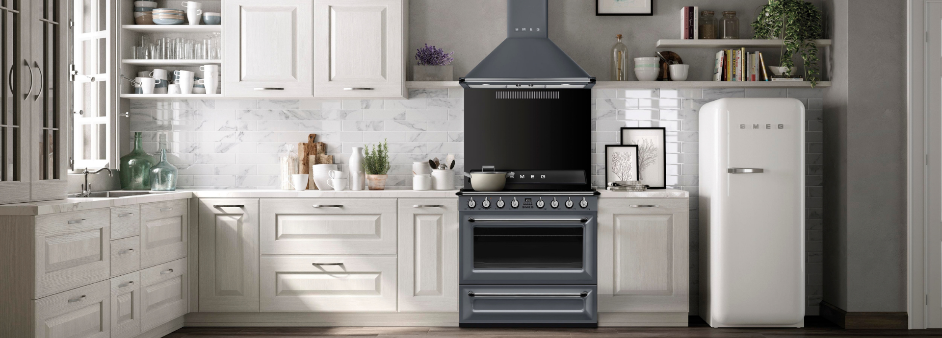 Piano de cuisson Smeg esthétique Victoria avec four intégré, finition gris ardoise, manettes de commande chromés, installé au centre d’une cuisine au style classique élégant. Au-dessus, une hotte décorative murale Smeg assortie avec finition gris ardoise et bandeau inférieur couleur inox surplombe l’espace de cuisson. À droite, un réfrigérateur Smeg style années 50 avec finition blanche brillante et poignées chromées apporte une touche rétro à l’ensemble. L’arrière-plan est composé de meubles de cuisine blancs, d’une crédence effet marbre clair et d’étagères ouvertes accueillant vaisselle, livres et éléments décoratifs, dans une ambiance lumineuse et intemporelle.