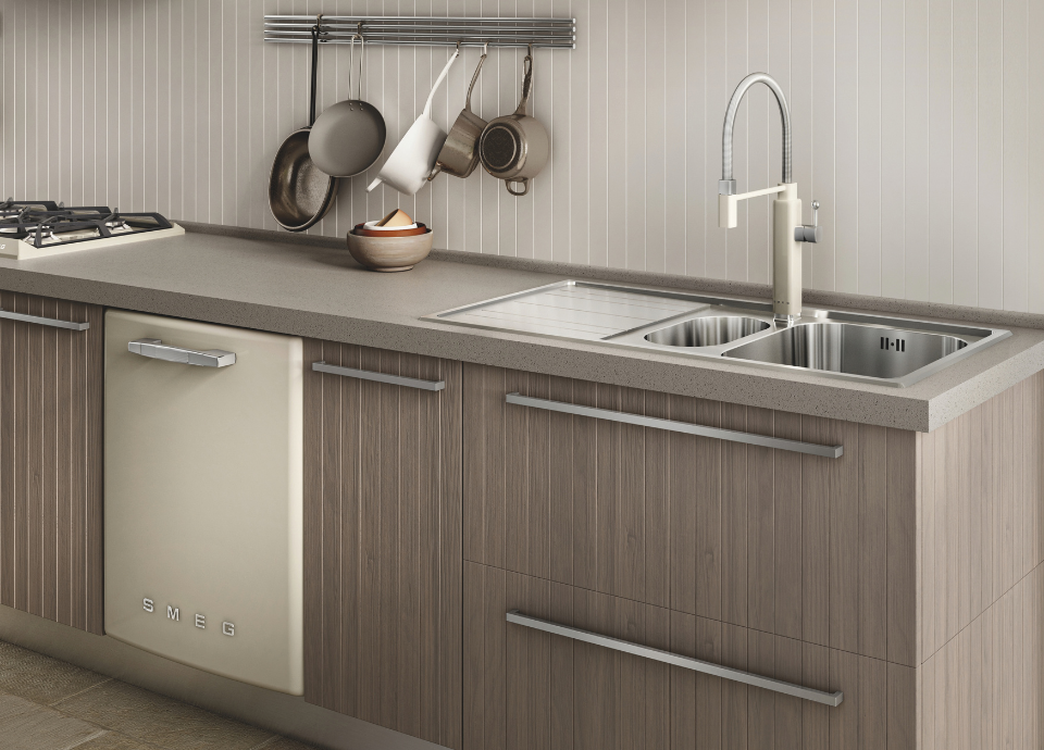 Lave-vaisselle encastrable Smeg avec façade crème et poignée horizontale en inox, intégré dans un linéaire de cuisine aux meubles effet bois et au plan de travail gris. À droite, un évier Smeg en couleur inox à deux cuves avec égouttoir intégré est équipé d'un mitigeur haut à douchette en couleur crème et inox. À l’arrière-plan, une crédence à lames verticales accueille une barre de suspension avec poêles, casseroles et tasses, tandis qu’une table de cuisson à gaz est visible à l’extrémité gauche du plan de travail.