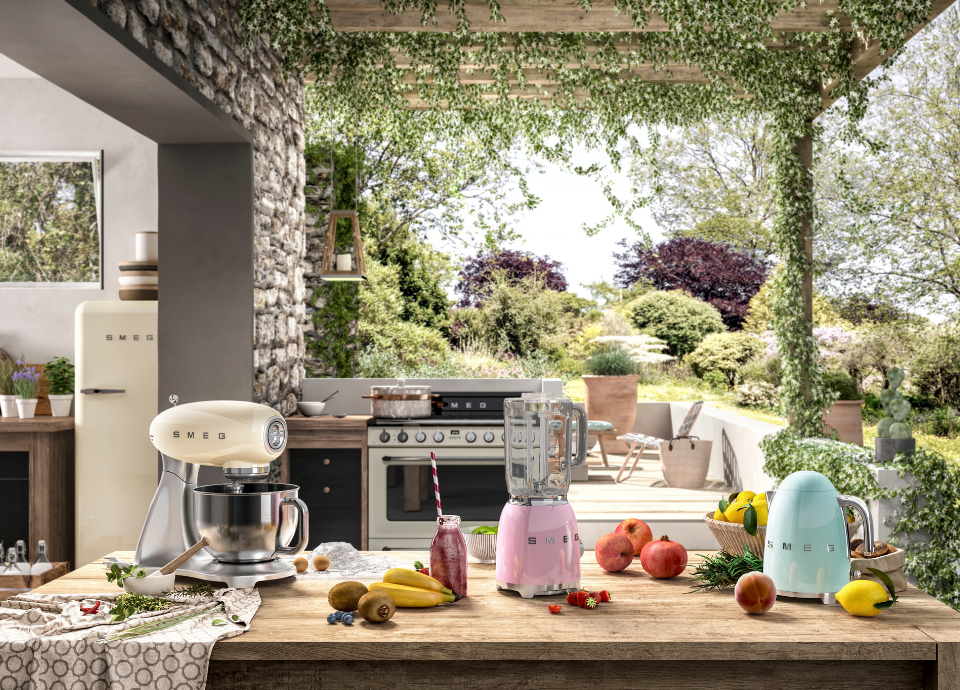 Petit électroménager Smeg composé d’un robot pâtissier crème avec bol en inox, d’un blender rose pastel avec poignée chromée et d’une bouilloire bleu pastel avec poignée chromée, disposés sur une table en bois entourée de fruits frais et d’herbes aromatiques. À l’arrière, une cuisinière Smeg est installée dans une cuisine d’été sous pergola végétalisée, avec vue sur un jardin paysager. Sur la gauche, un réfrigérateur crème Smeg style années 50 avec poignée chromée.