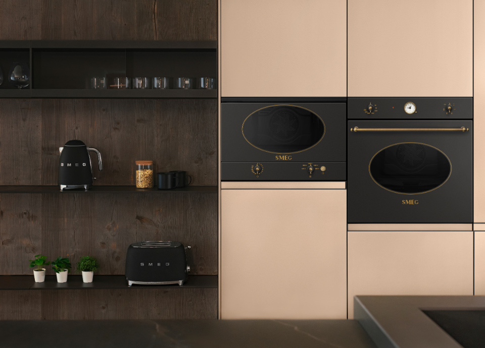 Four encastrable Smeg esthétique Coloniale avec façade anthracite, porte vitrée ovale, manettes de commande coloris laiton et horloge analogique centrale, intégré dans une colonne de cuisine aux façades beige clair. À ses côtés, un micro-ondes encastrable Smeg assorti reprend les codes esthétiques de la collection Coloniale avec finition anthracite, détails en laiton et fenêtre ovale caractéristique. Sur la gauche, une niche en bois foncé accueille une bouilloire et un grille-pain Smeg noirs, accompagnés d’accessoires de cuisine et de petites plantes décoratives.