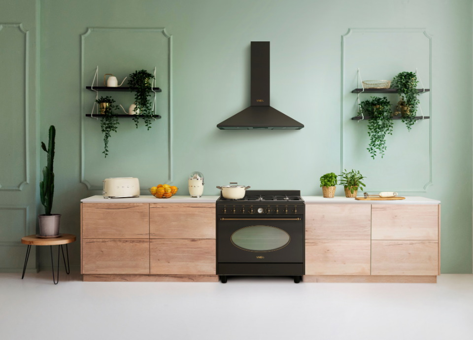 Hotte décorative murale Smeg esthétique Coloniale avec finition Anthracite, installée au-dessus d’un piano de cuisson Smeg esthétique Coloniale doté d’un four intégré, de manettes de commande en couleur laiton. L’ensemble est mis en scène dans une cuisine lumineuse aux meubles en bois clair et au mur vert sauge. Sur le plan de travail prennent place un grille-pain Smeg blanc, un presse-agrumes Smeg assorti, un saladier d’agrumes et plusieurs herbes aromatiques. L’arrière-plan est agrémenté d’étagères murales accueillant des objets décoratifs et des plantes retombantes, créant une ambiance chaleureuse mêlant design contemporain et inspiration classique.