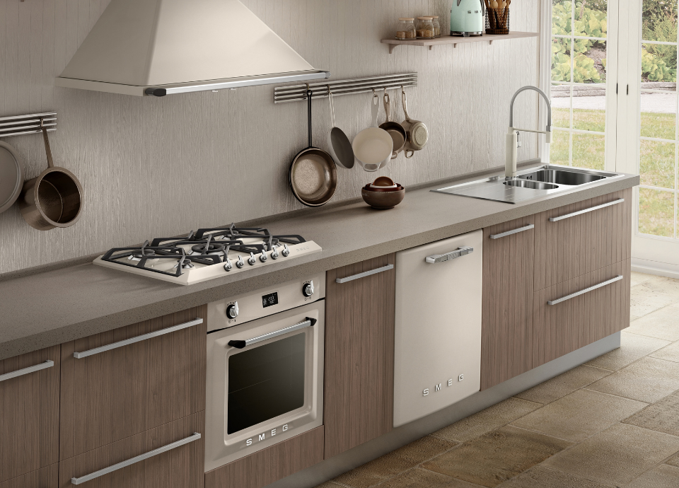 Plaque de cuisson à gaz Smeg esthétique Victoria en finition crème avec grilles noir, associée à une hotte décorative murale crème Smeg Victoria assortie dans une cuisine aux meubles effet bois et au plan de travail gris. À l’arrière-plan, des ustensiles de cuisson sont suspendus à une barre murale, tandis qu’un évier en inox avec un mitigeur, un four encastrable et un lave-vaisselle intégré Smeg complètent cet ensemble coordonné au design élégant et intemporel.