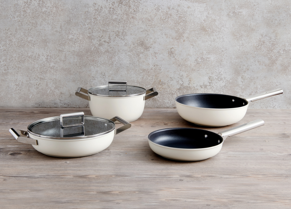 Batterie de cuisine Smeg de couleur crème composée de quatre ustensiles de cuisine. Elle comprend une poêles, une casseroles, une sauteuse et un wok. Les différents ustensiles sont disposés sur un plan de travail en bois. En arrière-plan, un mur aux teintes marron et noires