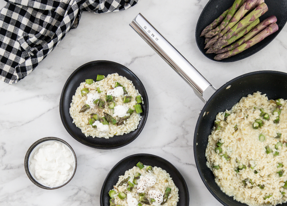 Wok Smeg noir présenté sur un plan de travail en marbre blanc. Il est placé sur la droite de l’image et contient un risotto aux asperges fraîchement préparé. Autour du wok, des assiettes avec les différents éléments de la préparation sont disposées sur le plan de travail. Un bouquet d’asperges vertes est placé dans la partie supérieure de l’image. En haut à gauche, un torchon à carreaux noir et blanc complète la mise en scène.