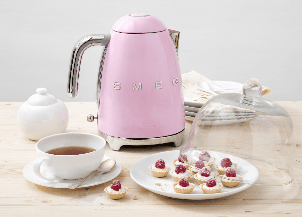 Bouilloire électrique SMEG 1,7L rose brillante au style années 50 avec poignée chromée, présentée sur une table avec une tasse de thé, des pâtisseries et de la vaisselle blanche.