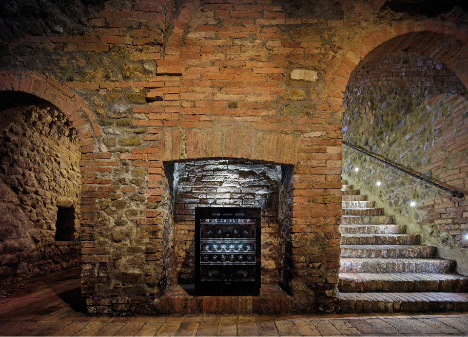 Cave à vin Smeg esthétique Dolce Stil Novo avec finition verre Noir Eclipse, intégrée au cœur d’une cave voûtée en pierre et briques apparentes. Dotée d’une porte en verre noir élégante, d’étagères de présentation illuminées, la cave à vin constitue l’élément principal de la composition. Installée dans une niche en briques anciennes, elle met en valeur les bouteilles grâce à un éclairage intérieur subtil. À l’arrière-plan, les murs en pierre naturelle, les arches en briques et l’escalier voûté éclairé créent une atmosphère authentique et prestigieuse.