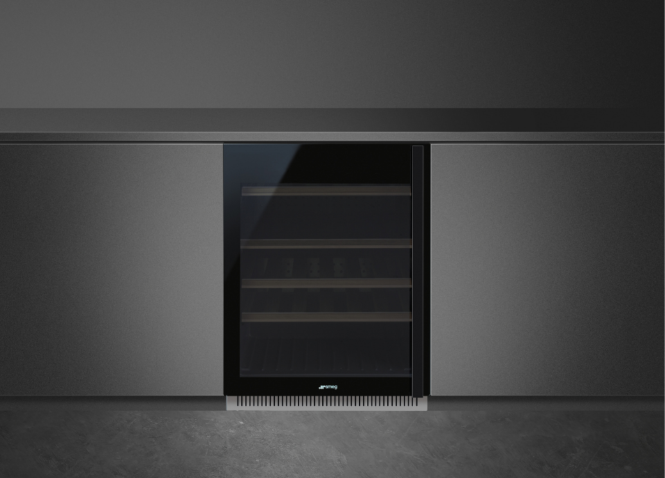 Cave à vin Smeg Dolce Stil Novo noire avec poignée rectangulaire intégrée sur toute la hauteur du côté droit de la porte. Une grille en inox est présente dans la partie inférieure. La vitre teintée laisse apparaître quatre tiroirs de rangement destinés aux bouteilles. La cave est encastrée dans un meuble gris placé devant un fond noir.
