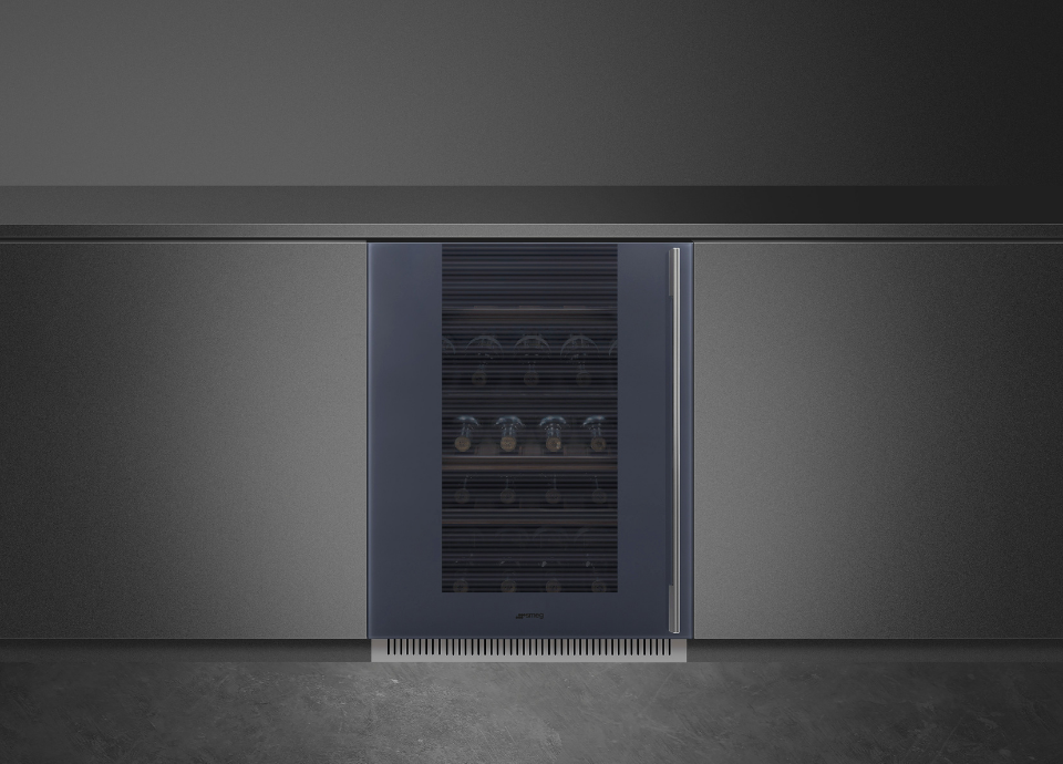 Cave à vin Smeg Linea bleu nuit et inox avec poignée inox arrondie intégrée sur toute la hauteur du côté droit de la porte. Une grille d’aération est visible dans la plinthe inférieure. La vitre semi-teintée noire à motifs rayés laisse apparaître les bouteilles à l’intérieur. La cave est encastrée dans un meuble gris devant un fond noir.