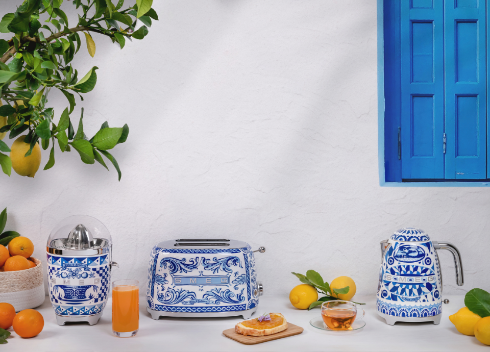 Collection Blu Mediterraneo Smeg composée d’un presse-agrumes, d’un grille-pain et d’une bouilloire, disposés sur un plan de travail clair dans un décor inspiré de la Méditerranée. Les appareils arborent une finition blanche et bleue ornée de motifs décoratifs emblématiques de la collection Blu Mediterraneo. Le grille-pain occupe le centre de la composition, entouré d’un presse-agrumes à gauche et d’une bouilloire à droite. À proximité, des oranges, des citrons, un verre de jus d’orange, une tasse de thé et une tartine agrémentée de marmelade apportent une touche gourmande à la scène. En arrière-plan, un citronnier et une fenêtre aux volets bleus.