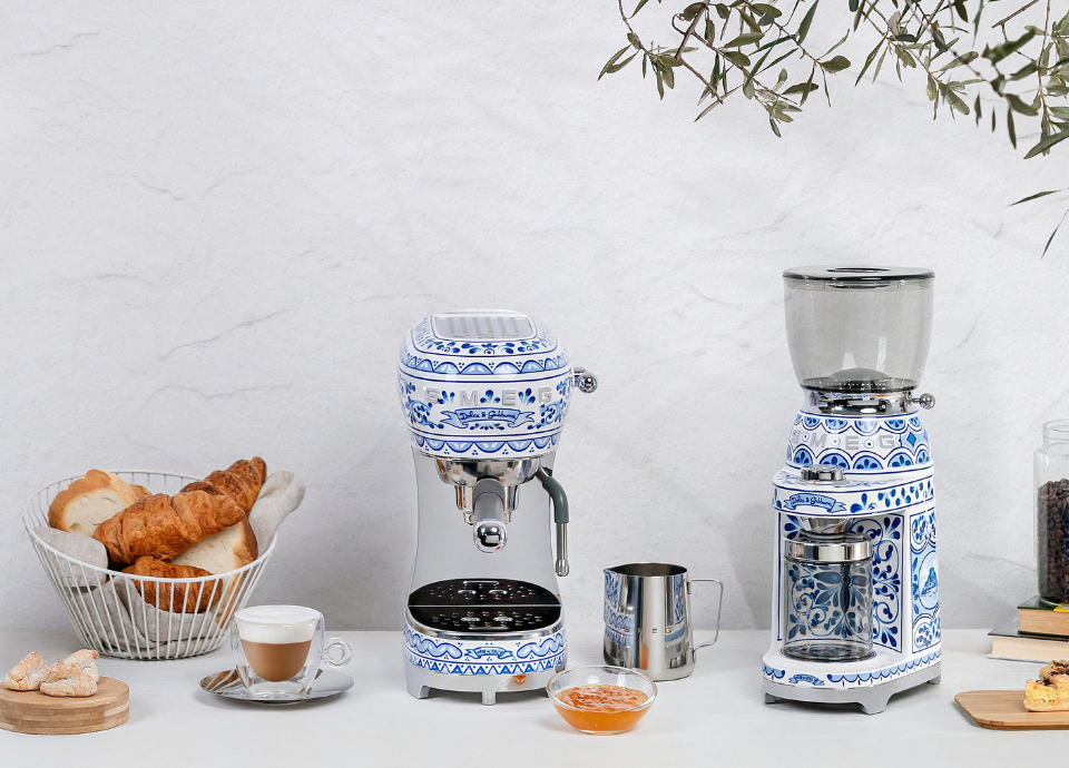 Collection Blu Mediterraneo Smeg composée d’une machine expresso et d’un broyeur à café, disposés sur un plan de travail clair dans un décor inspiré de la Méditerranée. Les appareils arborent une finition blanche et bleue ornée des motifs emblématiques de la collection Blu Mediterraneo. La machine expresso occupe le centre de la composition avec son porte-filtre chromé, sa buse vapeur et son plateau repose-tasses, tandis que le broyeur à café assorti est installé à ses côtés. À proximité, une tasse de café, un pichet à lait en inox, des viennoiseries et un bol de marmelade complètent la scène. En arrière-plan, des branches d’olivier et des grains de café.