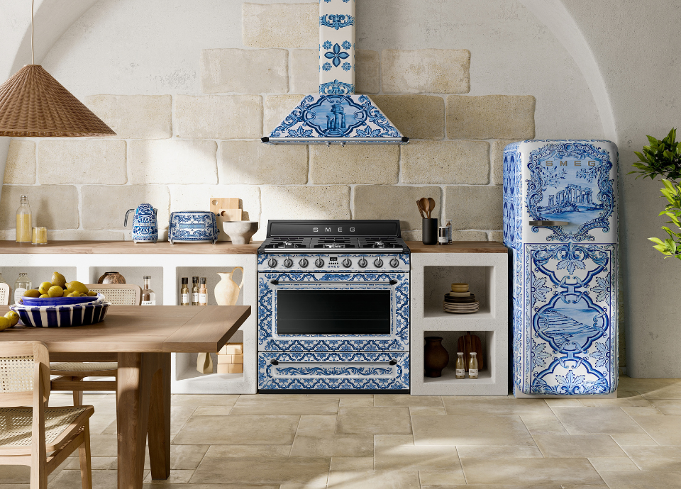 Collection Blu Mediterraneo Smeg composée d’un réfrigérateur FAB28, d’un centre de cuisson et d’une hotte décorative murale, installés dans une cuisine d’inspiration méditerranéenne. Les appareils arborent une finition blanche et bleue ornée des motifs emblématiques de la collection Blu Mediterraneo. Le centre de cuisson occupe le cœur de la composition avec sa large cavité de four, sa table de cuisson à gaz et ses manettes de commande coordonnées, tandis que la hotte assortie est installée au-dessus. À droite, le réfrigérateur FAB28 complète harmonieusement l’ensemble avec ses formes arrondies iconiques et ses décors artistiques inspirés des paysages côtiers italiens. Les meubles en maçonnerie, les éléments en bois naturel, les petits électroménagés de la même collection et les murs en pierre claire renforcent l’atmosphère chaleureuse