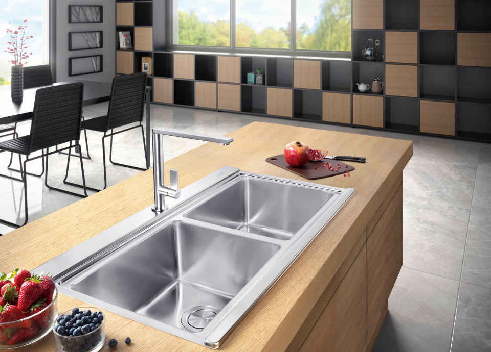 Évier Smeg esthétique Linéa à deux cuves en couleur inox, intégré dans un îlot central en bois clair au sein d’une cuisine contemporaine. Doté de bacs spacieux aux angles épurés et d’un égouttoir intégré, l’évier est associé à un mitigeur chromé au design moderne. À proximité, des fruits rouges dans un bol en verre ainsi qu’une grenade posée sur une planche à découper apportent une touche de couleur au plan de travail. En arrière-plan, de larges bibliothèques aux finitions bois et noir, une table de repas avec chaises contemporaines et de grandes baies vitrées ouvertes sur un paysage verdoyant