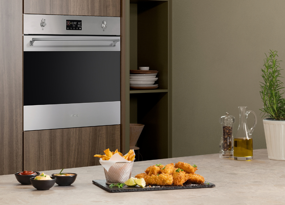 Four encastrable Smeg esthétique Classica avec finition inox et verre Noir Eclipse, intégré dans une colonne de meubles à l’aspect bois naturel. Doté d’une large poignée tubulaire en couleur inox, de manettes de commande métalliques et d’un afficheur numérique central, le four constitue l’élément principal de la composition. Au premier plan, un plat de pièces panées accompagné de quartiers de citron, de frites servies dans un panier métallique et de sauces est présenté sur un plan de travail effet pierre clair. À proximité, une huilière en verre, un moulin à poivre transparent et une plante aromatique apportent une touche décorative. En arrière-plan, une niche de rangement ouverte accueille une pile d’assiettes aux tons clairs