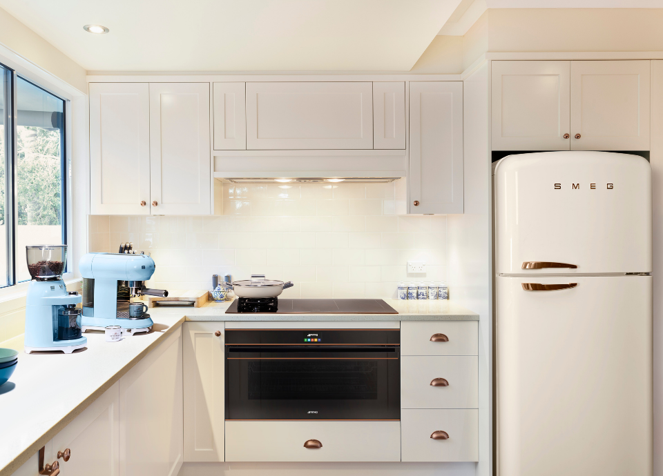 Four encastrable Smeg esthétique Dolce Stil Novo avec finition noir, détails cuivrés et verre céramique Noir Eclipse, intégré sous une table de cuisson dans une cuisine lumineuse aux façades blanches. Doté d’un bandeau de commande élégant, d’un affichage numérique discret, le four constitue l’élément principal de la composition. Au-dessus, une table de cuisson intégrée est installée sous une hotte encastrée discrète. À proximité, une machine à espresso bleu pastel, un moulin à café assorti et plusieurs accessoires décoratifs apportent une touche chaleureuse à l’espace. En arrière-plan, un réfrigérateur iconique Smeg à pose libre de couleur crème complète l’aménagement.