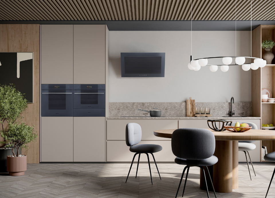 Fours encastrables Smeg esthétique Linéa, finition verre clair standard couleur Neptune Grey, intégrés en colonne dans un ensemble de meubles aux lignes minimalistes. Les fours se distinguent par leurs poignées horizontales en inox et leurs affichages numériques discrets, constituant l’élément central de la composition. À proximité, une hotte murale assortie en finition Neptune Grey est installée au-dessus du plan de travail. L’espace est complété par un mobilier aux tons clairs, un plan de travail effet pierre, un évier avec mitigeur noir et une table à manger ronde entourée de chaises contemporaines. En arrière-plan, des étagères décoratives, des accessoires de cuisine et un luminaire suspendu aux globes opalins renforcent l’atmosphère élégante, chaleureuse et contemporaine de la pièce.