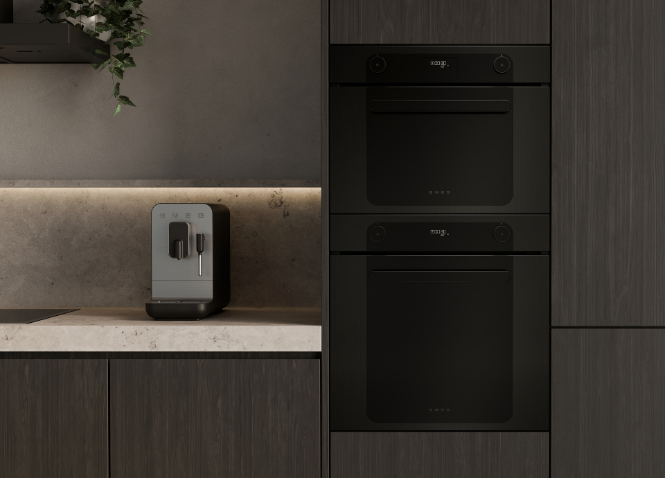 Fours encastrables Smeg esthétique Musa avec finition noire et verre Noir Eclipse, installés en colonne dans une cuisine contemporaine aux façades effet bois foncé. Dotés d’un design minimaliste, d’écrans numériques discrets et de commandes tactiles intégrées, ils s’intègrent harmonieusement dans l’architecture de la cuisine. À gauche, une machine à café automatique Smeg gris avec buse vapeur est disposée sur un plan de travail clair devant une crédence effet béton. L’ensemble est sublimé par un éclairage indirect sous meuble et des éléments végétaux suspendus qui renforcent l’atmosphère élégante et raffinée de l’espace.
