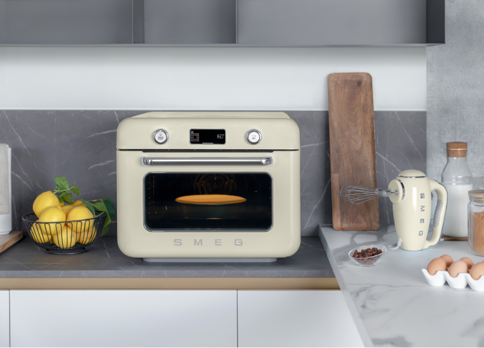 Mini four combiné vapeur pose libre Smeg couleur crème au style années 50, installé dans une cuisine moderne en marbre gris. Le four est doté d’une longue poignée chromée sur la porte et d’une porte vitrée noire située sous l’écran de réglage. L’écran au dessus affiche les différents modes de cuisson et est encadré de deux boutons de commande blancs avec un contour chromé. Le logo Smeg chromé est placé sous la porte vitrée. À gauche, un panier en métal noir contient des citrons. À droite, un batteur électrique Smeg couleur crème est accompagné d’une planche, d’œufs et de fruits secs.