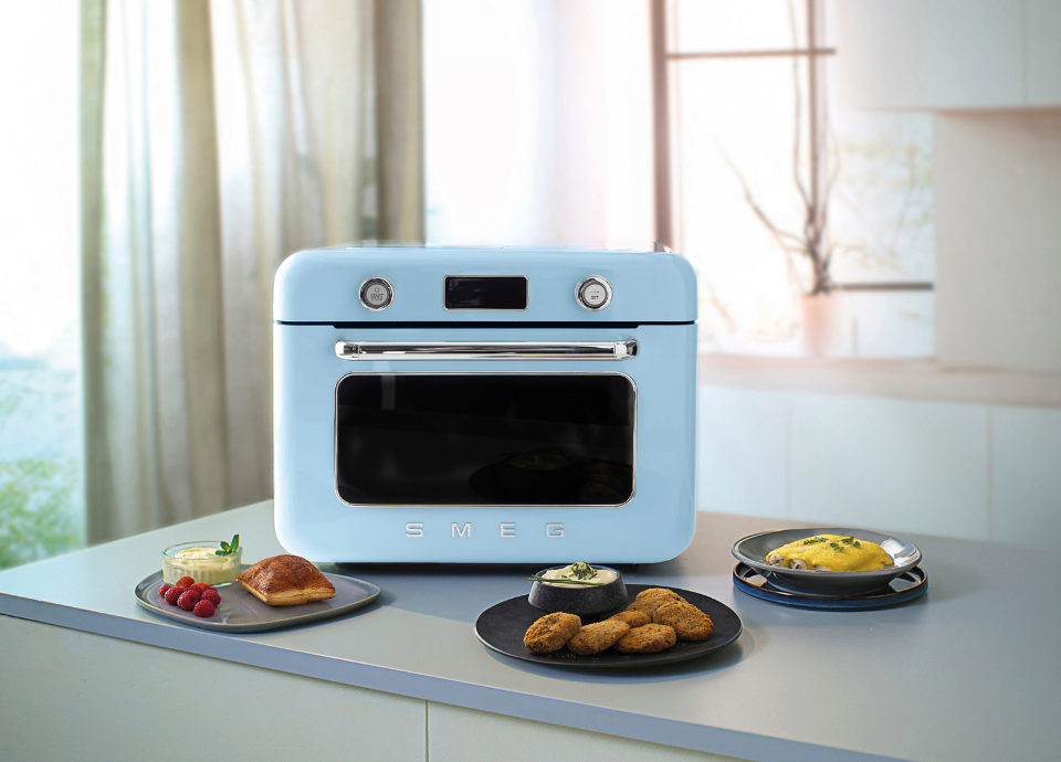 Mini four combiné vapeur pose libre Smeg couleur bleu au style années 50, installé dans une cuisine moderne beige avec un plan de travail gris. Le four est doté d’une longue poignée chromée sur la porte et d’une porte vitrée noire située sous l’écran de réglage. L’écran affiche les différents modes de cuisson et est encadré de deux boutons de commande chromés, avec un contour chromé et une partie supérieure blanche. Le logo Smeg chromé est placé sous la porte vitrée. Au premier plan, trois assiettes sont disposées devant le four : la première contient un dessert à base de crème et de framboises, la deuxième des nuggets accompagnés d’une sauce aux herbes et la troisième une purée de butternut avec des champignons. À l’arrière-plan, des fenêtres avec des rideaux marron clair apportent de la lumière à la cuisine.
