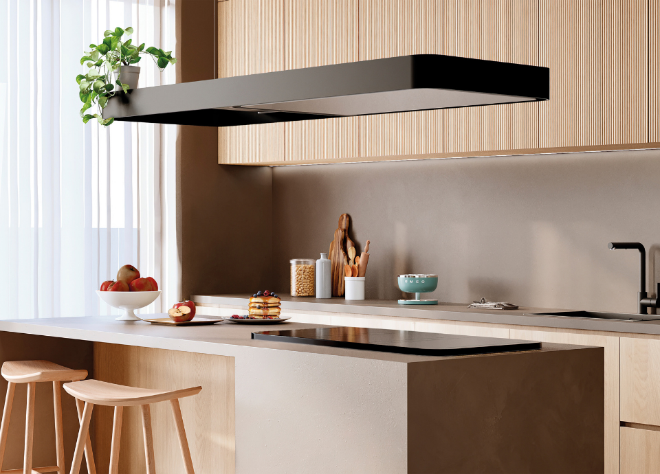 Hotte décorative îlot central Smeg, esthétique Isola, avec finition noir mat et design rectangulaire suspendu aux lignes épurées, installée au-dessus d’un vaste îlot de cuisine contemporaine. La hotte constitue l’élément central de la composition et intègre un éclairage discret sous sa structure. En dessous, une table de cuisson en verre noir est encastrée dans le plan de travail clair de l’îlot. À proximité, un bol de fruits, une assiette de pancakes et des assises en bois clair. En arrière-plan, des meubles aux finitions bois naturel, un évier avec mitigeur noir, des accessoires de cuisine dont la balance Smeg vert emeraude.
