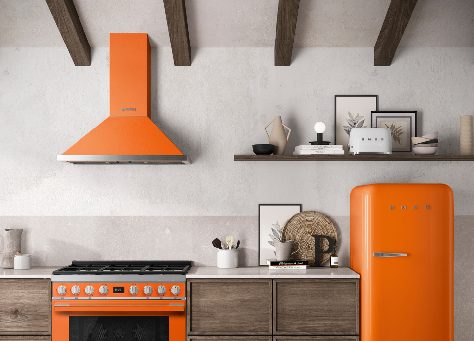 Hotte décorative murale Smeg esthétique Portofino de couleur orange, installée comme élément central de la composition au-dessus d’un piano de cuisson assorti. Dotée d’un design aux lignes franches et de finitions en inox, la hotte apporte une touche vive et contemporaine à la cuisine. En dessous, un piano de cuisson Smeg Portofino de couleur orange complète harmonieusement l’ensemble avec sa table de cuisson à gaz et ses manettes métalliques. À proximité, un réfrigérateur iconique Smeg assorti, des étagères en bois et plusieurs objets décoratifs