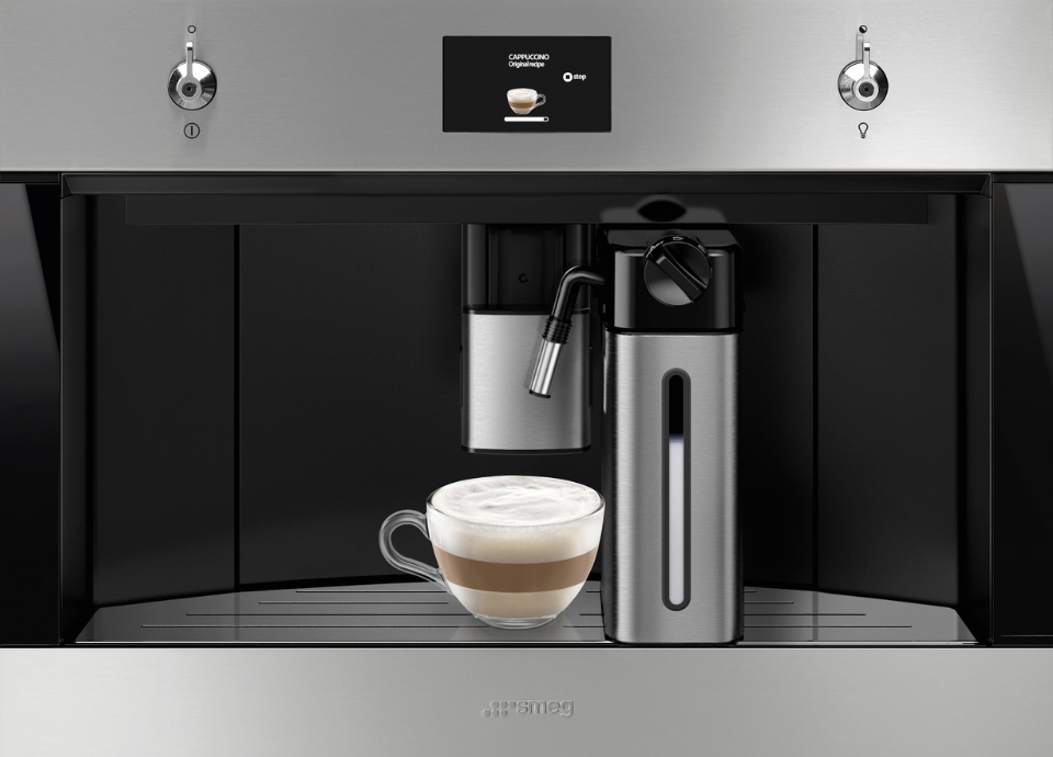 Machine à café encastrable Smeg esthétique Classica en couleur inox et verre Noir Eclipse, intégrée dans un meuble de cuisine. Dotée de manettes de commande métalliques, d’un écran numérique central et d’un système de préparation automatique, la machine constitue l’élément principal de la composition. Au centre de l’espace de service, une tasse en verre contenant un cappuccino à la mousse onctueuse est placée sous le bec verseur, à côté d’une buse à vapeur. L’intérieur de la cavité, habillé de finitions noires et d’un plateau repose-tasse en couleur inox, met en valeur les détails techniques de l’appareil.