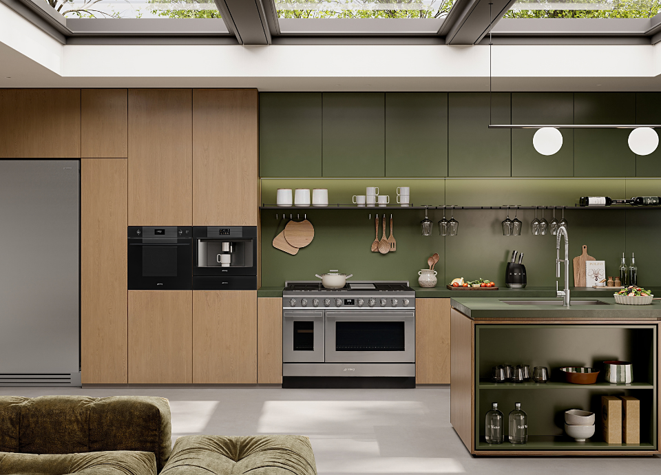 Machine à café encastrable Smeg esthétique Linéa avec finition verre noir standard, intégrée dans une colonne de meubles en bois naturel au sein d’une cuisine contemporaine. Dotée d’un écran de commande discret, d’un bec verseur central et d’un habillage en verre élégant, la machine à café constitue l’élément principal de la composition. À ses côtés, un four encastrable coordonné complète l’ensemble avec des lignes épurées et une façade assortie. En arrière-plan, un piano de cuisson en inox, une crédence vert olive, des accessoires suspendus et un îlot central avec évier créent un espace chaleureux et fonctionnel. Une grande verrière en toiture apporte une abondante lumière naturelle.