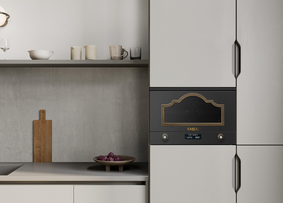 Micro-ondes avec gril encastrable Smeg esthétique Cortina, doté d’une façade anthracite, d’un encadrement de porte coloris laiton antique, d’un logo doré et de manettes assorties, intégré dans une colonne de cuisine aux façades beige clair. À gauche, une étagère murale accueille de la vaisselle et des accessoires décoratifs, tandis qu’un plan de travail clair met en valeur une planche en bois et un saladier de fruits.