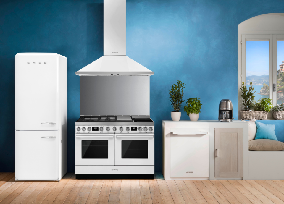 Piano de cuisson Smeg esthétique Portofino de couleur blanche, largeur 120 cm, installé au centre d’une cuisine lumineuse au style méditerranéen. Doté d’une large table de cuisson à gaz, de deux cavités de cuisson, de manettes métalliques et d’afficheurs numériques intégrés, le piano constitue l’élément principal de la composition. Au-dessus, une hotte décorative murale blanche assortie complète harmonieusement l’ensemble. À proximité, un réfrigérateur iconique Smeg blanc, des plantes aromatiques et une machine à café s’intègrent dans un décor aux murs bleu profond et aux matériaux naturels.