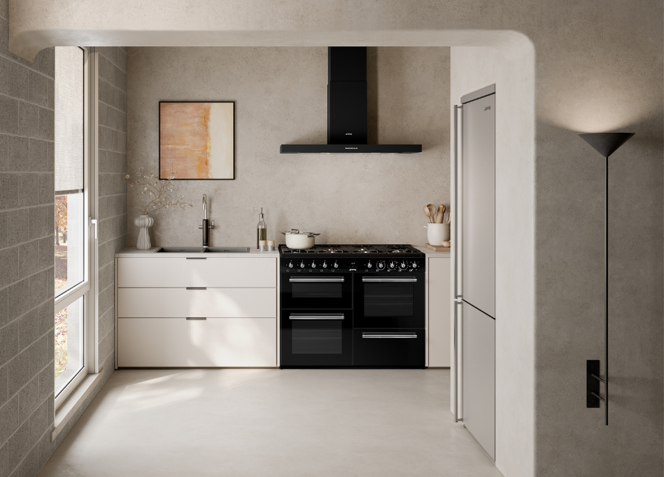 Piano de cuisson Smeg esthétique Classica avec finition noir et détails chromés, installé au centre d’une cuisine contemporaine aux tons clairs. Doté de plusieurs cavités de cuisson, de manettes de commande métalliques et d’une large table de cuisson à gaz, le piano constitue l’élément principal de la composition. Au-dessus, une hotte murale Smeg noire aux lignes épurées est installée contre un mur à l’aspect minéral. À proximité, un plan de travail clair accueille une casserole, des ustensiles de cuisine et une bouteille d’huile. À l’arrière-plan, un réfrigérateur pose libre de couleur blanche, un évier avec mitigeur assorti et de grandes ouvertures laissant entrer la lumière