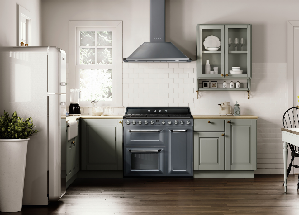 Piano de cuisson Smeg esthétique Victoria de 100 cm avec triple four intégré, finition gris ardoise, table de cuisson en émail noir et manettes de commande en inox, installé au centre d’une cuisine au style classique contemporain. Au-dessus, une hotte murale Smeg en couleur gris ardoise est fixée devant une crédence en carreaux métro blancs. Sur son côté droite, des meubles vert sauge avec vitrine qui exposent de la vaisselle et des verres, tandis que sur la gauche un blender est disposé sur le plan de travail. Sur le devant du plan de travail, un réfrigérateur combiné Smeg style années 50 avec compartiment congélateur supérieur et réfrigérateur inférieur complète cet ensemble aux inspirations rétro et élégantes.