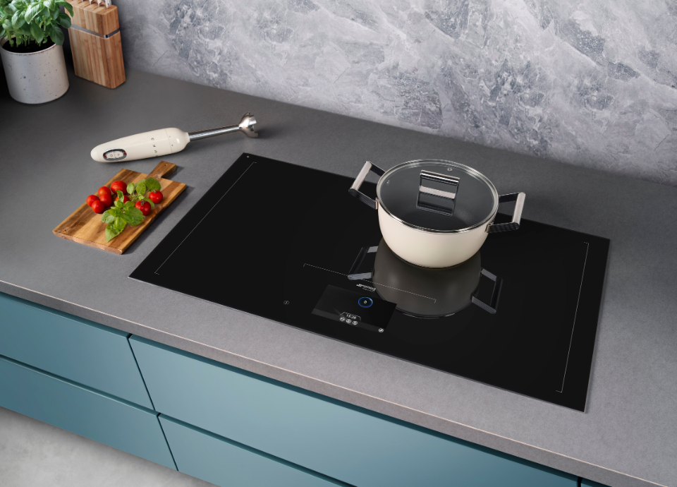Table de cuisson à induction Smeg intégrée dans un plan de travail gris effet pierre, avec surface vitrocéramique noire et commandes tactiles frontales. Une casserole Smeg crème avec couvercle en verre est positionnée sur une zone de cuisson. À gauche, une planche en bois présente des tomates cerises et des feuilles de basilic fraîches, tandis qu’un mixeur plongeant Smeg crème et des accessoires de cuisine complètent l’environnement. L’ensemble s’intègre dans une cuisine moderne aux meubles bleu mat et au revêtement mural effet marbre gris