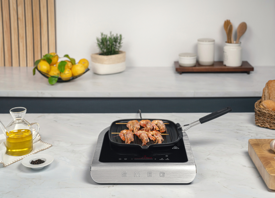Plaque à induction portable Smeg blanche avec finition inox et surface vitrocéramique noire, installée sur un plan de travail en marbre clair dans une cuisine contemporaine. Une poêle-gril carrée est posée sur la zone de cuisson et contient plusieurs brochettes de crevettes en cours de préparation. À proximité, une bouteille d’huile d’olive, un petit récipient de poivre, une coupe de citrons frais, des ustensiles de cuisine et une corbeille à pain complètent l’espace, dans un décor moderne aux lignes épurées.