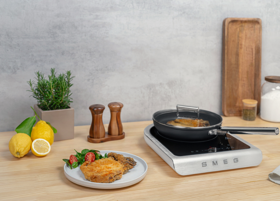 Plaque à induction portable blanche Smeg avec finition inox et surface vitrocéramique noire, posée sur un plan de travail en bois clair. Une poêle noire avec couvercle contient une préparation en cours de cuisson. Au premier plan, une assiette présente une escalope panée accompagnée de tomates cerises et de salade. À l’arrière, des citrons, un pot d’herbes aromatiques, des moulins à sel et poivre en bois ainsi que des accessoires de cuisine sont disposés dans un décor de cuisine moderne.