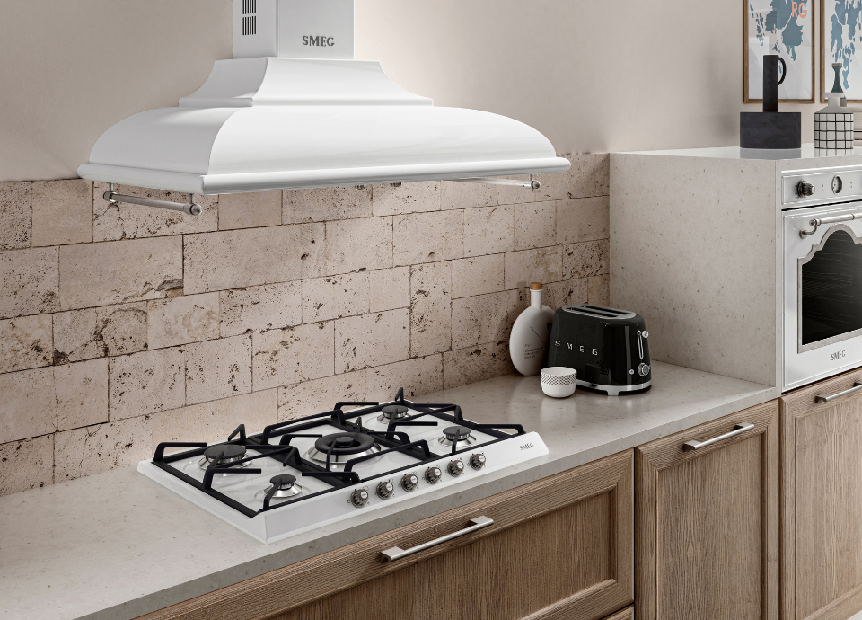 Plaques de cuisson à gaz Smeg esthétique Cortina avec finition crème, grilles noir et manettes de commande métalliques en façade, intégrée dans un plan de travail clair au sein d’une cuisine aux façades en bois naturel. Au-dessus, une hotte décorative murale Smeg de style traditionnel avec finition crème et barre porte-ustensiles en inox surplombe l’espace de cuisson. À droite, un grille-pain Smeg noir est disposé sur le plan de travail aux côtés d’accessoires décoratifs, tandis qu’un four encastrable Smeg assorti complète l’aménagement. L’ensemble est mis en valeur par une crédence en pierre naturelle.