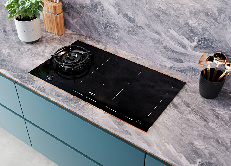 Table de cuisson mixte Smeg esthétique Dolce Stil Novo avec finition verre céramique Noir Eclipse et détails cuivrés, intégrée dans un plan de travail à l’aspect marbre gris. Dotée d’un brûleur à gaz central et de zones de cuisson à induction sérigraphiées. À proximité, un pot d’ustensiles aux reflets cuivrés, un moulin en bois et une plante aromatique apportent une touche décorative raffinée. L’ensemble est installé dans une cuisine aux façades bleu grisé et aux surfaces minérales.