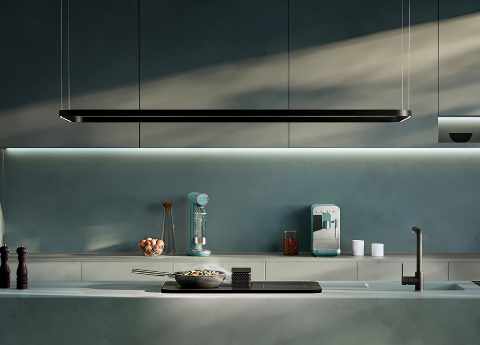 Table de cuisson à induction Smeg avec hotte intégrée esthétique Isola, dotée d’une finition en verre noir et de commandes tactiles frontales discrètes, installée sur un plan de travail clair au sein d’une cuisine contemporaine. Le système d’aspiration est intégré directement au centre de la plaque de cuisson, à proximité d’une poêle utilisée pour la préparation des aliments. Au-dessus de l’îlot, une suspension lumineuse linéaire noire apporte un éclairage élégant à l’espace. À l’arrière-plan, une machine à café et une machine à eau gazeuse Smeg vert emeraude. On retrouve également es accessoires de cuisine, un évier avec mitigeur noir et des façades aux lignes épurées.