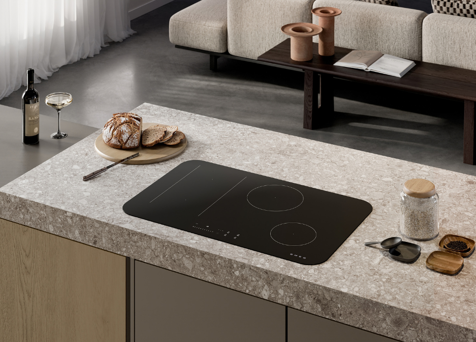 Plaque de cuisson à induction Smeg esthétique Isola avec finition en verre noir, intégrée dans un vaste îlot de cuisine à l’aspect pierre claire. La table est équipée de zones de cuisson sérigraphiées et de commandes tactiles frontales discrètes. À proximité, un pain de campagne tranché est disposé sur une planche en bois, accompagné d’une bouteille de vin et d’un verre. En arrière-plan, un espace de vie contemporain avec canapé beige, table basse en bois foncé et objets décoratifs crée une ambiance élégante et chaleureuse.