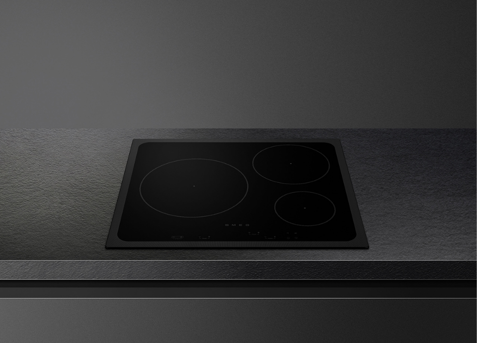 Plaque à induction Smeg esthétique Musa avec verre Noir Eclipse et finition noire, intégrée dans un plan de travail effet pierre foncée. Son design épuré à commandes tactiles et ses zones de cuisson discrètement dessinées soulignent le caractère contemporain et raffiné de la collection Musa.