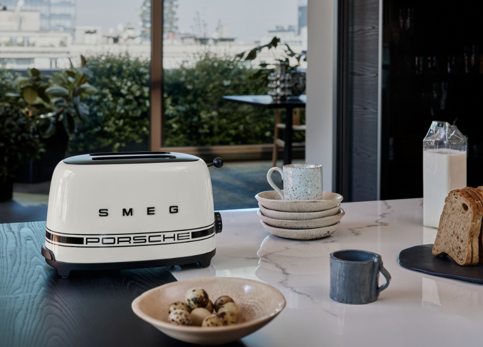 Grille-pain 2 tranches SMEG X Porsche collection Blanc Carrara au style années 50 avec poignée chromée, présenté sur une table entouré d’assiettes, de tasses et d’aliments.